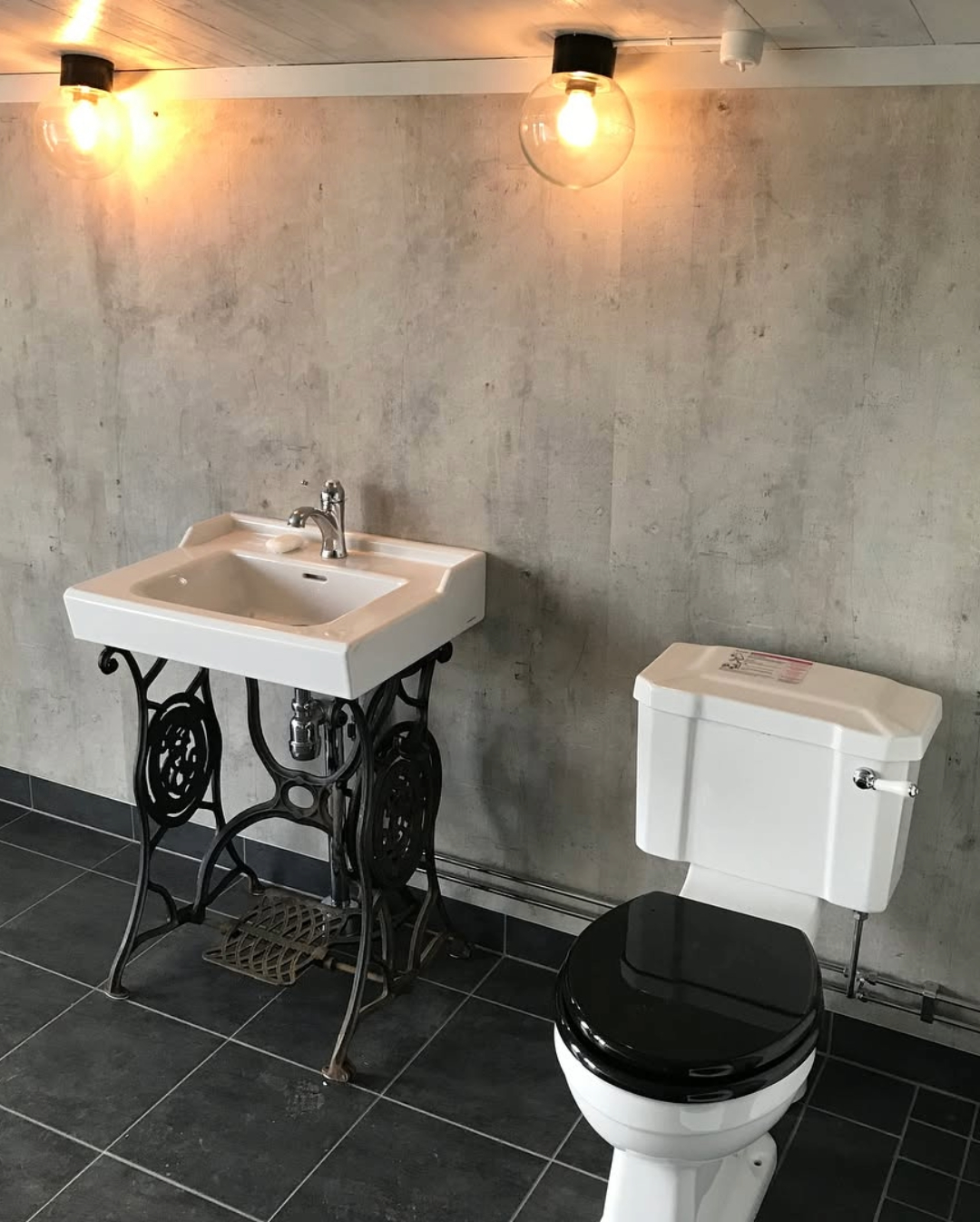 badrum med handfat på gammalt symaskinsunderrede, toalett med högspolande cistern och betongliknande vägg